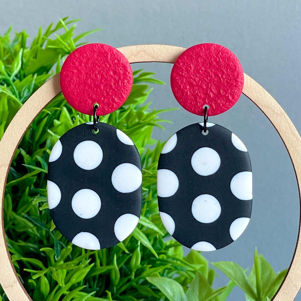 Pair of earrings with red tops and black and white polka dot bottoms on a green leafy background.