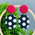 Pair of earrings with red tops and black and white polka dot bottoms on a green leafy background.
