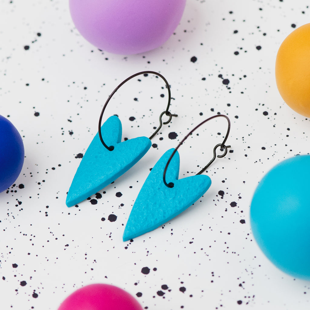 A pair of handmade turquoise heart-shaped hoop earrings on black hoops, displayed on a white speckled surface surrounded by colourful polymer clay balls.