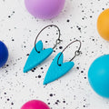 A pair of handmade turquoise heart-shaped hoop earrings on black hoops, displayed on a white speckled surface surrounded by colourful polymer clay balls.