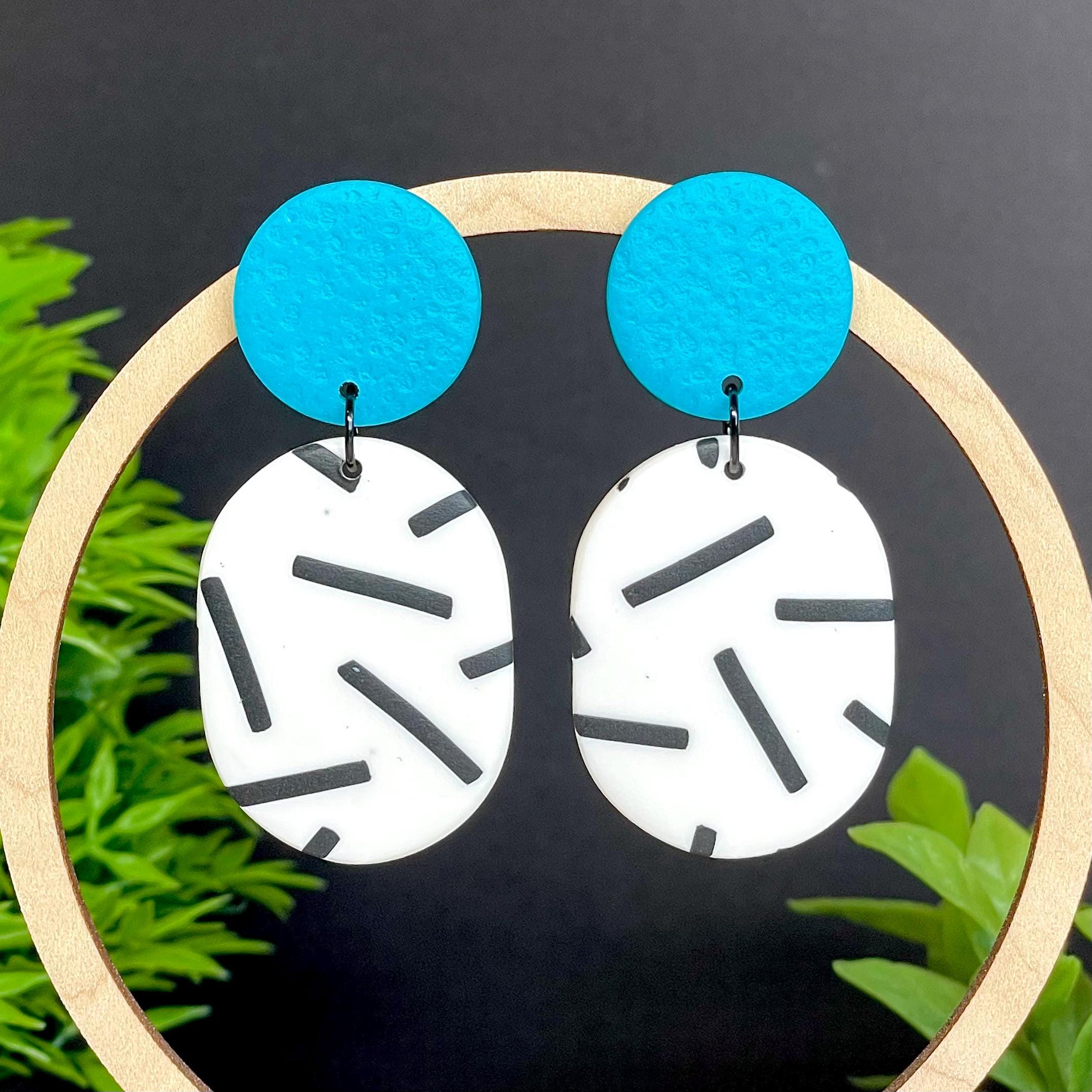 Earrings with turquoise tops and monochrome sprinkle oval bottoms, displayed on a dark background with greenery.